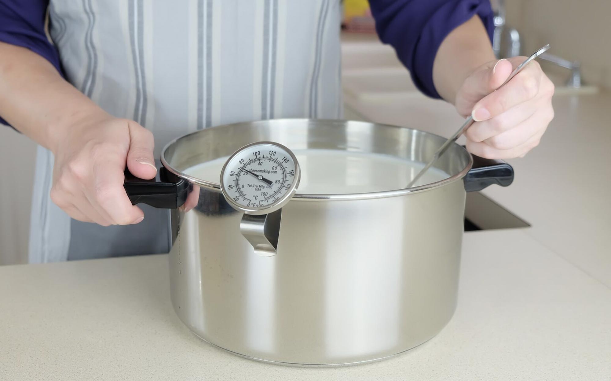 Person stirring milk in a stainless steel pot for cheese making with a thermometer restign on the edge.