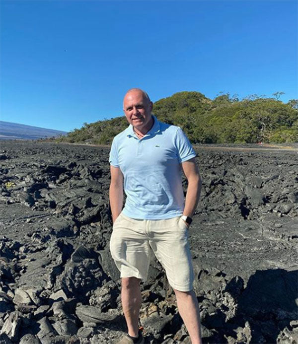 Cheesemaker Jamie Ryer, a white man with a short, military-style haircut stands on a hardened field of lava. He is wearing a light blue Lacoste polo, khaki shorts, and has his hands in his pockets.