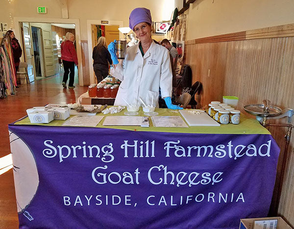 Woman standing behind her cheese stand with goat and chef hat on