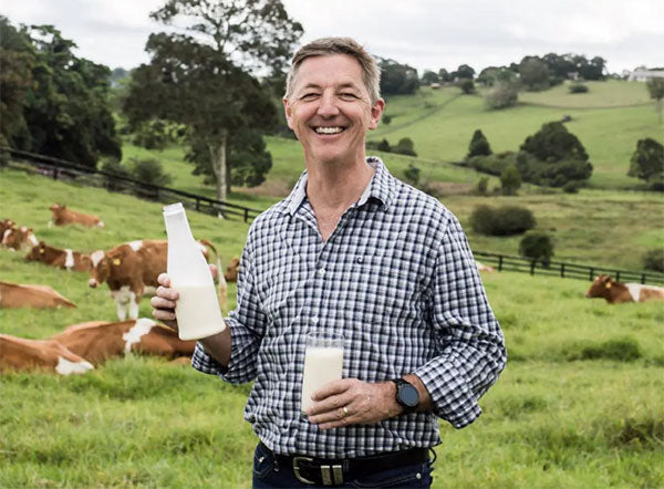 A man stands in a field of cows holding a glass of milk in one hand and a bottle of milk in the other