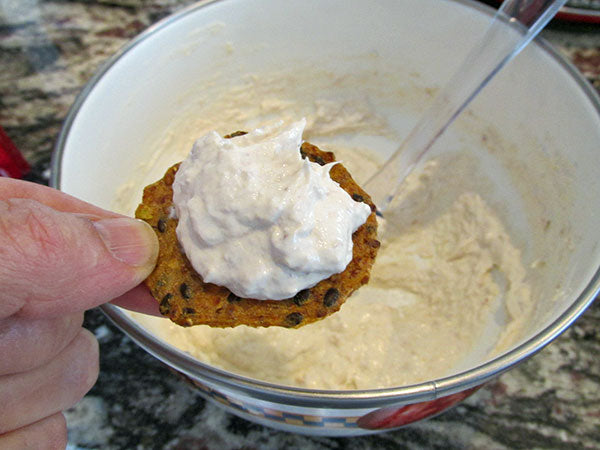 A hand holds a type of flat cracker topped with homemade smoked tuna cream cheese over a bowl of the same mixture.