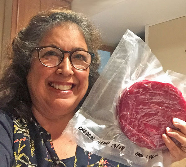 A woman in dark rimmed round glasses selfies while holding a shrink-wrapped wheel of homemade cheese