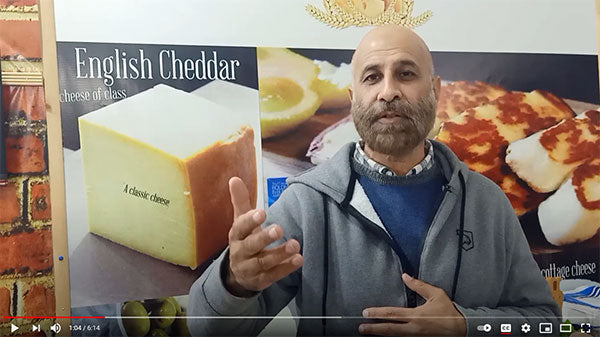 A screenshot of a YouTube video with a light-skinned man, bald, with a light brown beard, gray jacket over a blue sweater and collared shirt appears to be explaining something. There is a poster of cheese behind him.