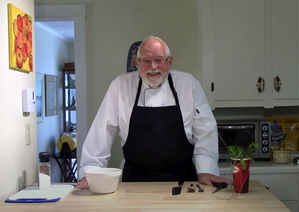 A white man with glasses, white receeding hair, and a white beard and mustache stands at a wooden countertop in a kitchen wearing a white chef's coat and black apron. There are kitchen cabinets behind him.