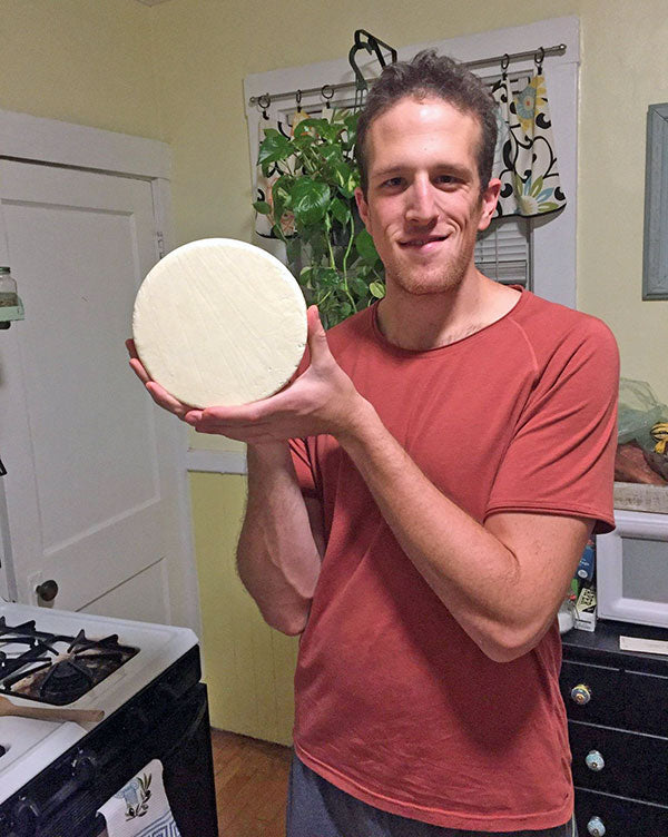 A cheese maker stands in a kitchen in front of a stove holding a wheel of cheese