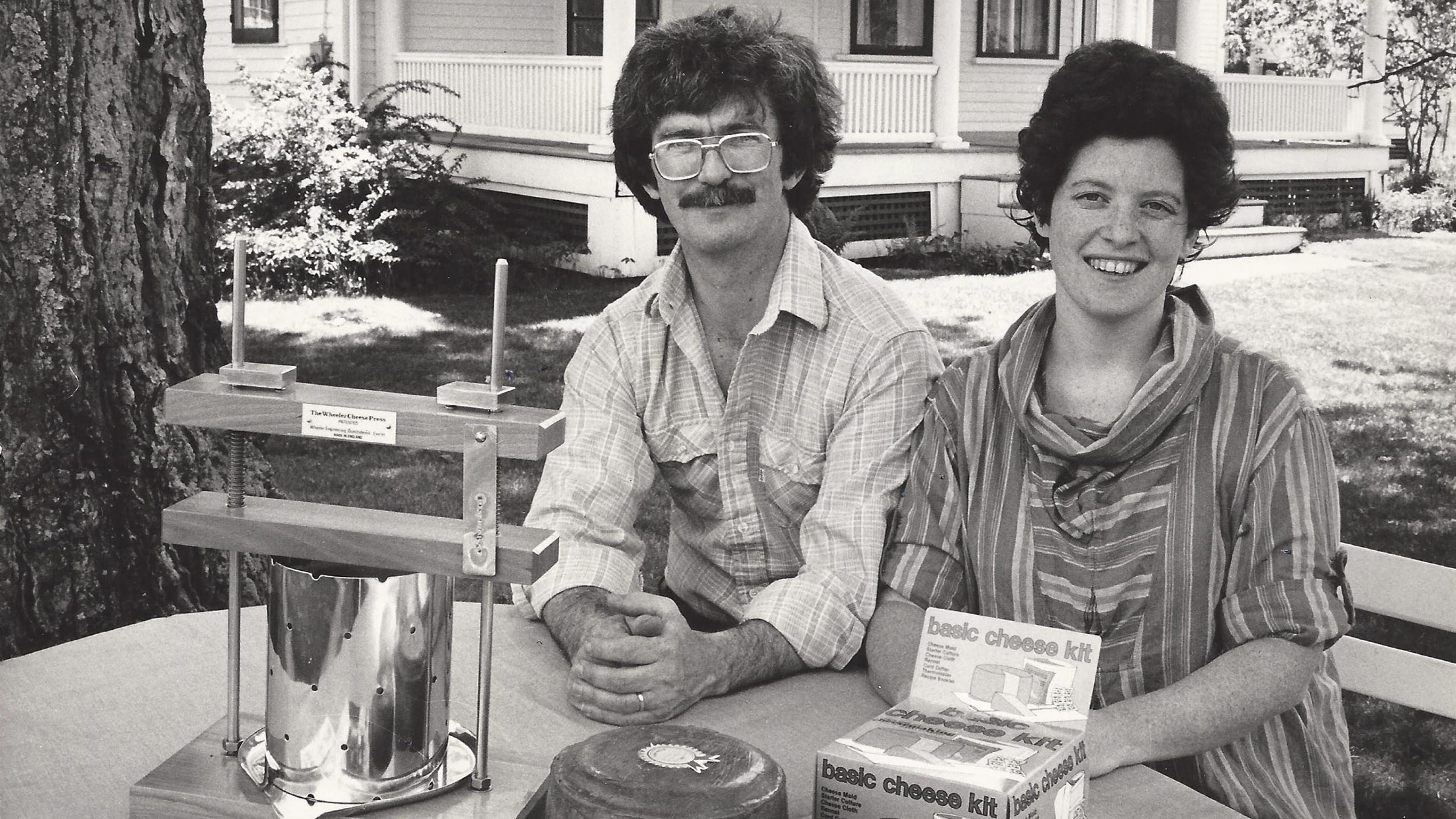 Black and white photo of Robert and Ricki Carroll, founders of New England Cheesemaking Supply Company, sitting outside at a table with a cheese press, waxed homemade cheese and a cheese kit